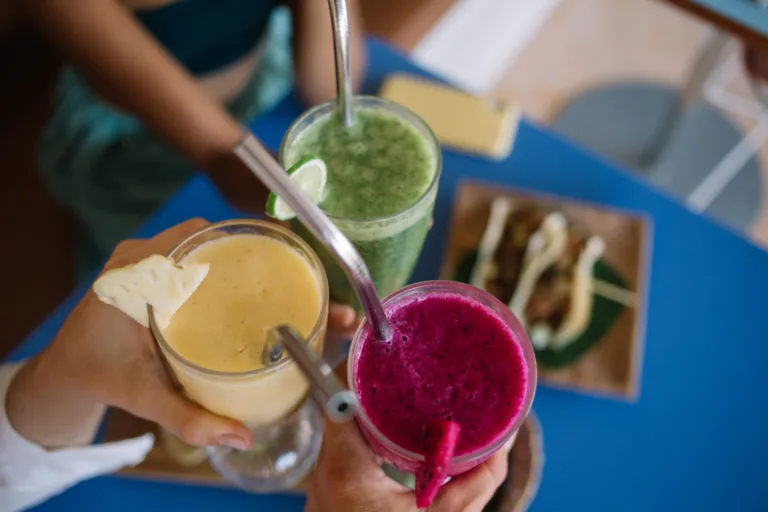 Cropped image of cafeteria customers holding smoothie cocktails and gesturing cheers during friendly meeting in bistro, selective focus on colorful milkshakes with mixed ripe fruits and fresh veggies