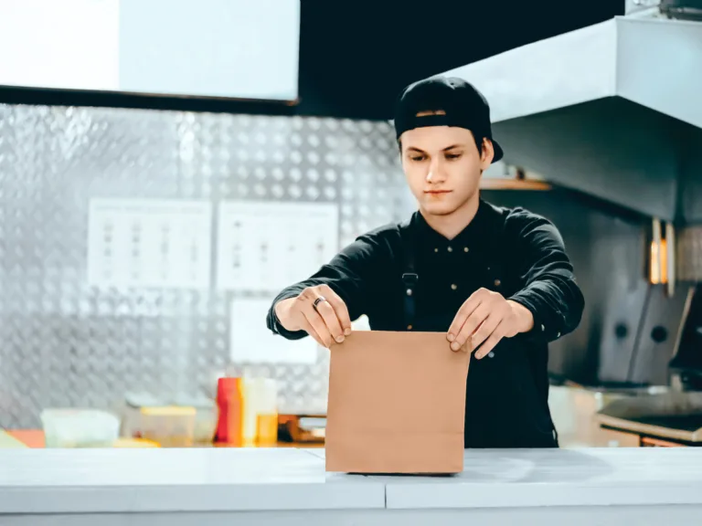 Waiter in black uniform is packing takeaway food in a paper bag while standing behind the counter