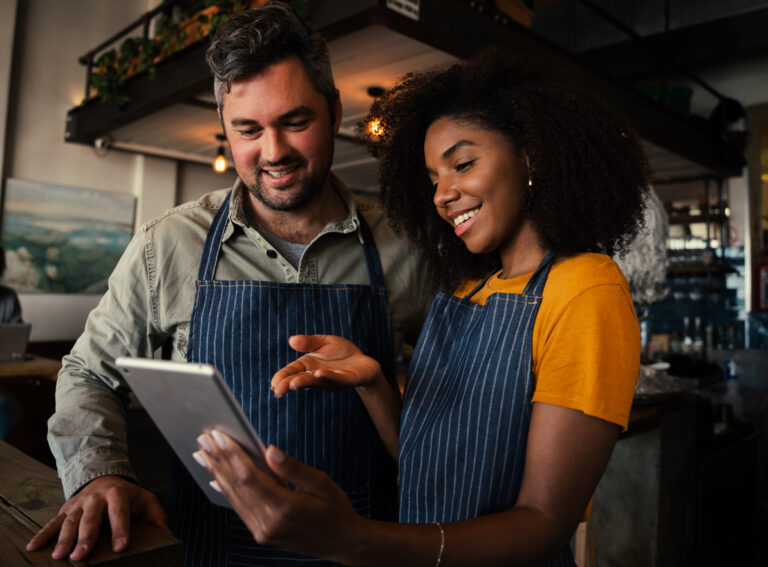 Smiling white male and ethnic female colleagues discussing business plans while scrolling on a digital tablet standing in trendy coffee shop. High quality photo
