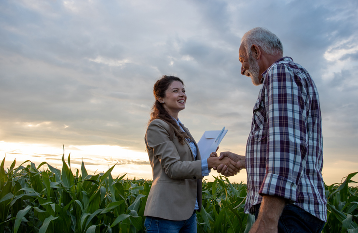 Senior man farmer shaking hands with young pretty woman with notebook in corn field. Insurance in agribusiness concept
