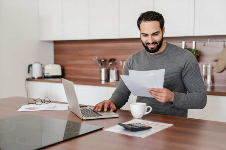Busy concentrated Middle Eastern man freelancer student counting calculating costs, expenses, income, rental taxes, doing paperwork in the kitchen using laptop. Remote work occupation, finance concept