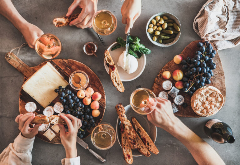 Flat-lay of human hands drinking wine eating fresh cheese fruits bread wine snacks over grey concrete table background, top view. Gathering, celebrating, wine tasting and party concept
