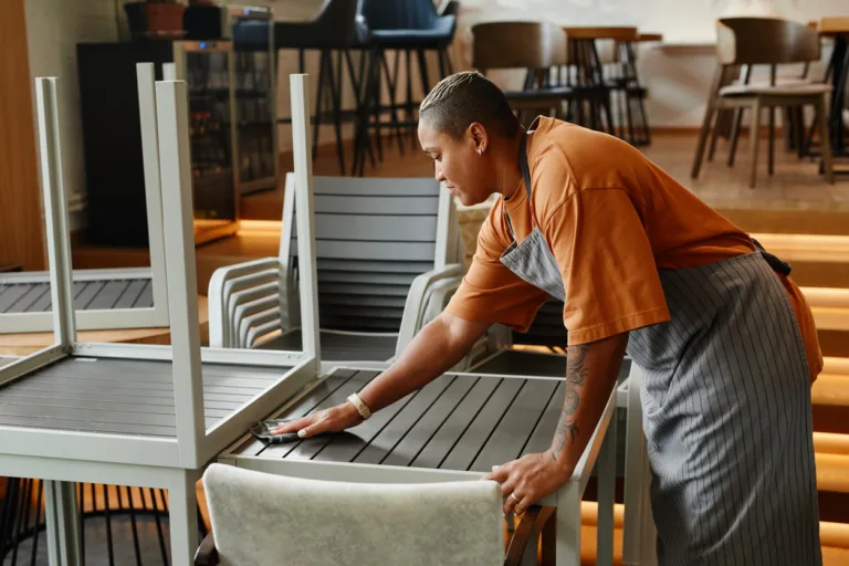 Young adult African American woman starting workday in modern cafe preparing tables turning them over and wiping dust