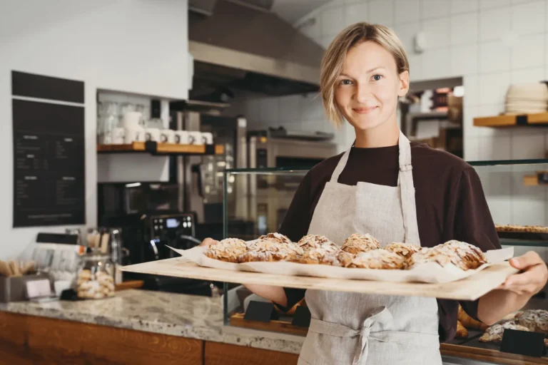 Welcoming female baker holding freshly baked almond croissants in background of bakery. Young smiling seller woman in cafe or coffee shop. Bistro owner indoors. Sustainable local small business.
