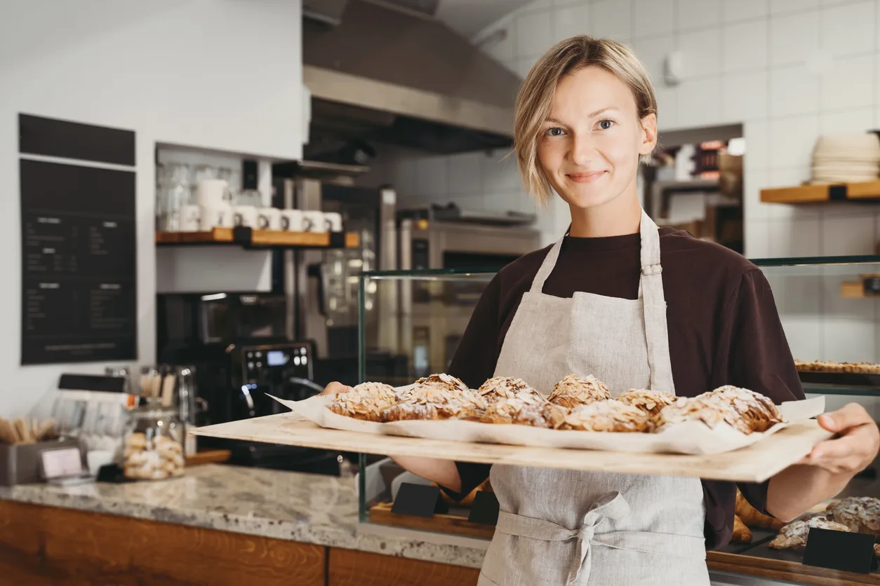 Welcoming female baker holding freshly baked almond croissants in background of bakery. Young smiling seller woman in cafe or coffee shop. Bistro owner indoors. Sustainable local small business.