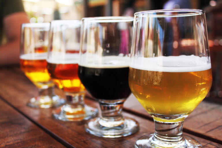 Flight of four beers lined up on a wooden table, with selective focus on the first beer. Photo taken outdoors with natural lighting; beers range in color from light to very dark. Background out of focus.