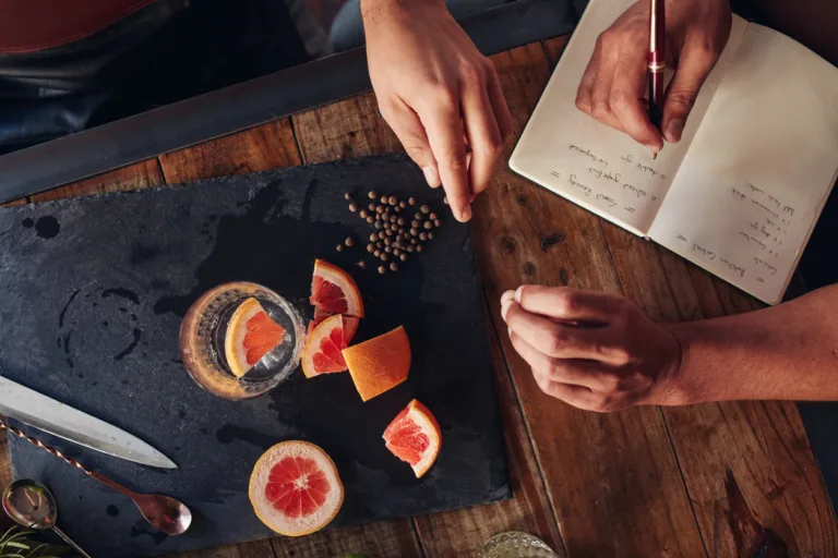 Top view of ingredients for making a cocktail with bartender taking notes. Cut grapefruit and black peppercorn on chopping board.