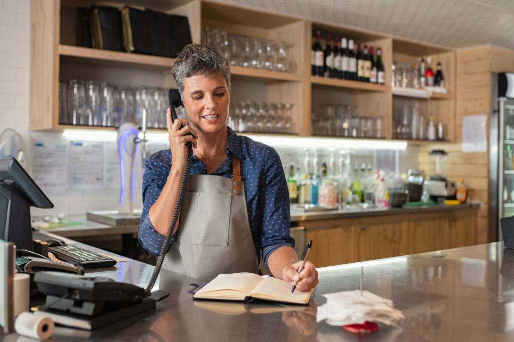 Barista woman taking orders over the phone at counter. Mature woman in grey apron writing on customer book while talking over phone in restaurant counter. How to reduce food waste in restaurants