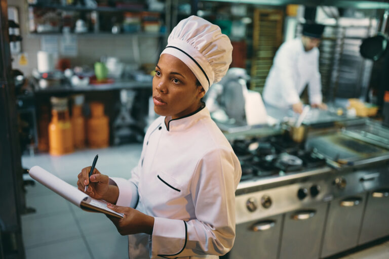 African American female chef taking notes while examining stock of groceries at restaurant's kitchen. restaurant equipment maintenance checklist