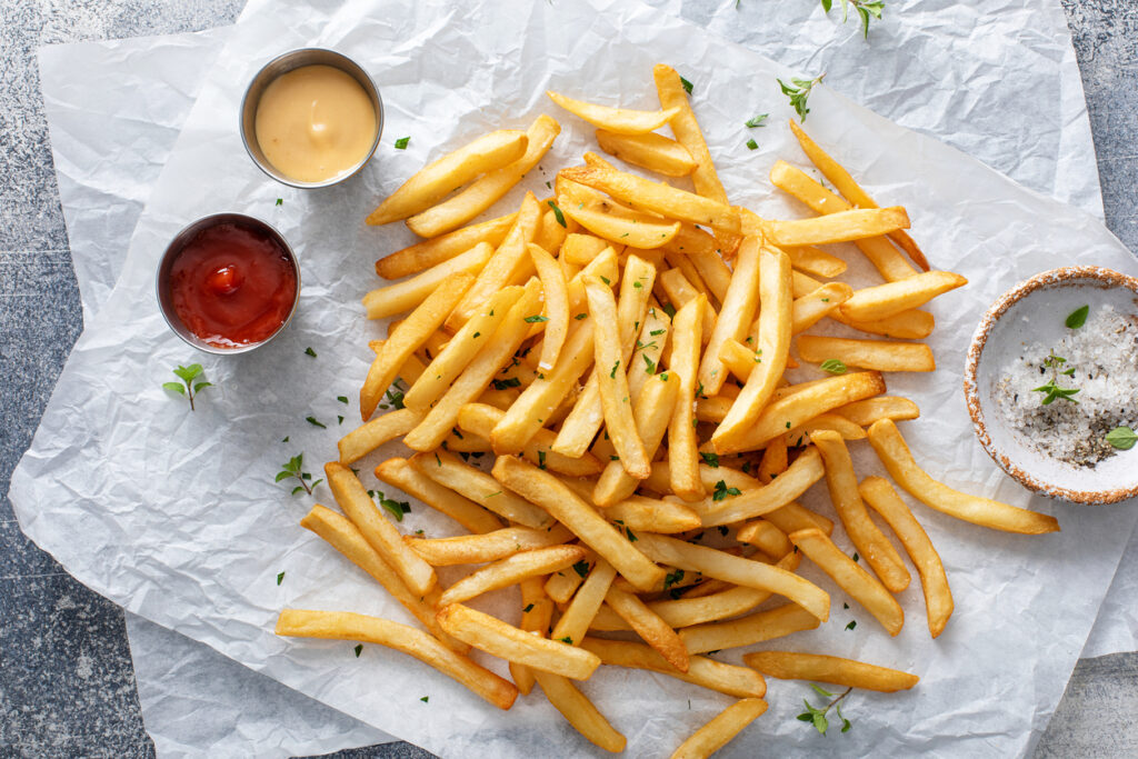 French fries on parchment paper with ketchup and mustard. A bowl of salt and herbs nearby.