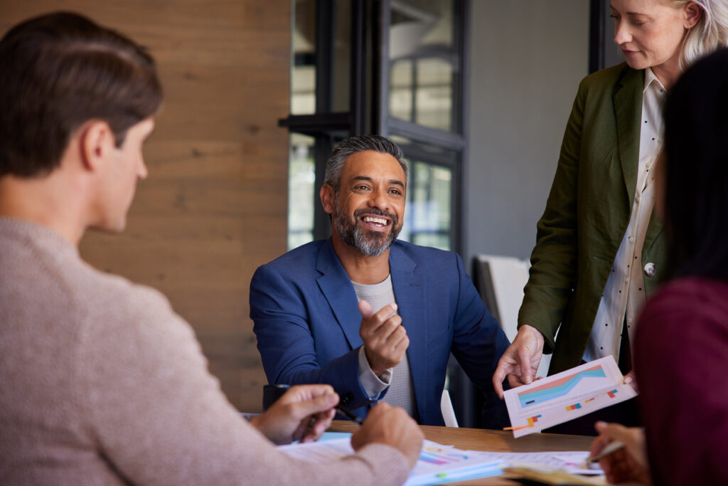 What is procurement supply chain management? Business people share printouts of charts and discuss at conference table. Smiling man in center.
