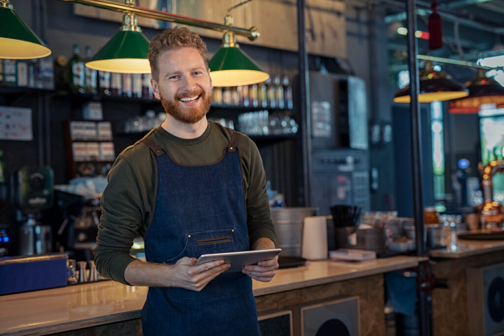Successful small business owner using digital tablet and looking at camera. Happy smiling waiter wearing apron and holding digital tablet ready to take order. Portrait of young entrepreneur of coffee shop standing at counter with copy space.