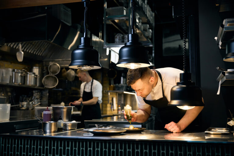 Caucasian male chef pouring sweet sauce from bottle on cheesecakes with kiwi slices on plate at table in restaurant. Concept of delicious healthy eating