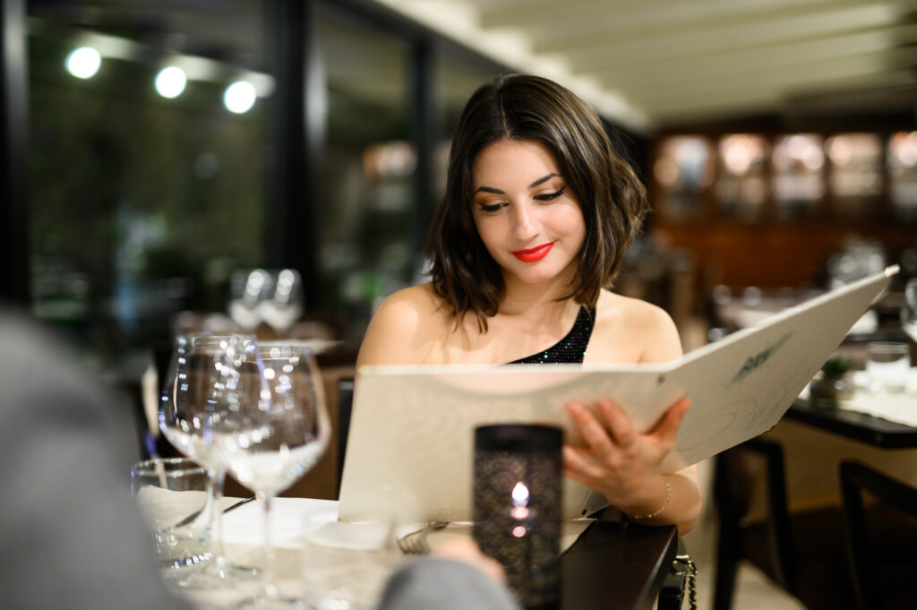 Young woman with red lipstick reads a fancy restaurant menu, choosing dinner in a luxurious setting