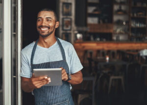 Young smiling african entrepreneur standing at the entrance of his trendy cafe holding digital tablet looking away