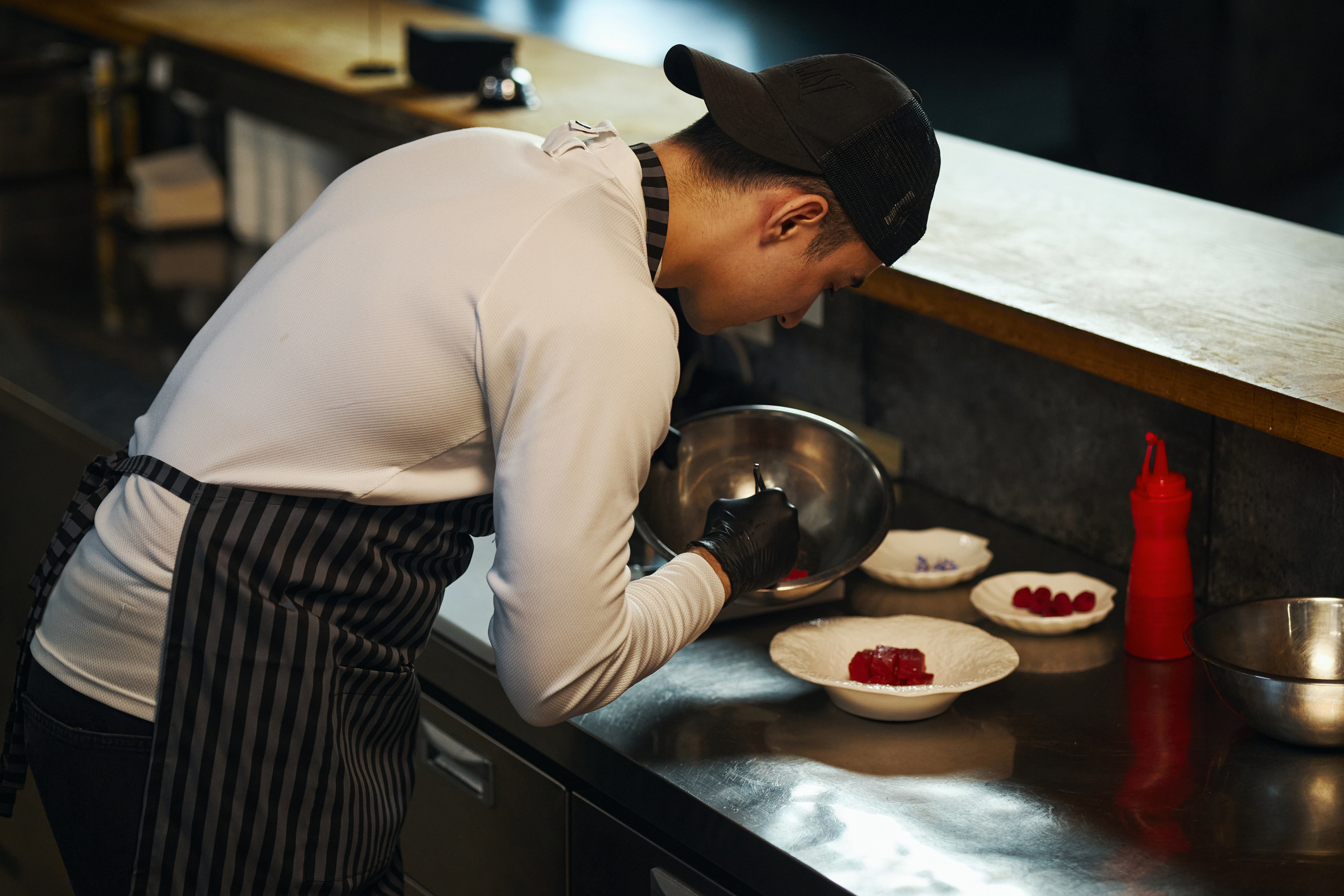 Young adult Asian man preparing dessert in professional kitchen, wearing apron and cap, carefully plating food with metal bowl, arranging ingredients on plate