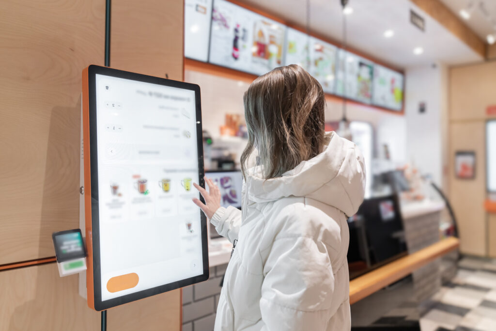 A young woman interacts with a digital kiosk in a stylish caf, selecting items for her order, A woman in a white jacket chooses drinks and orders food for dessert in a restaurant or pizzeria coffee shop through a touchscreen screen on her own. The restaurant menu is displayed on the screen in the terminal.