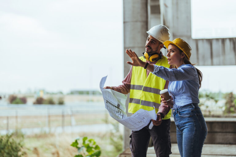 Young woman architect explaining blueprint to supervisor wearing safety vest at construction site. Mid adult contractor holding blueprint and understanding manager vision at construction site. foodservice purchasing