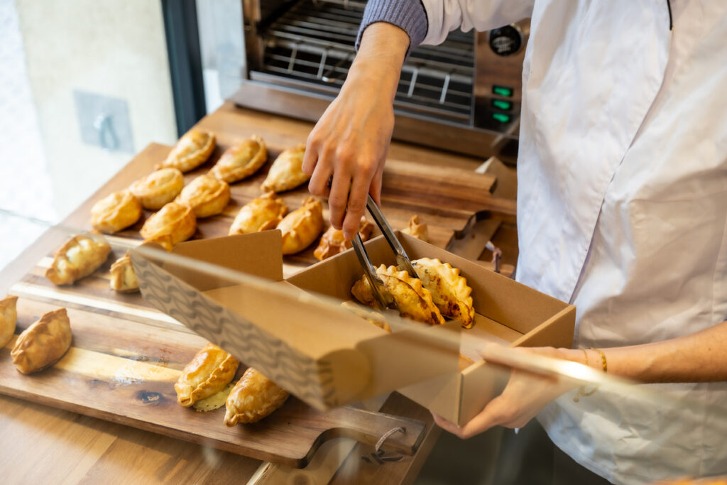 Chef's hands using tongs to carefully place traditional savory empanadas from a wooden board into kraft paper takeaway boxes, showcasing fresh food service